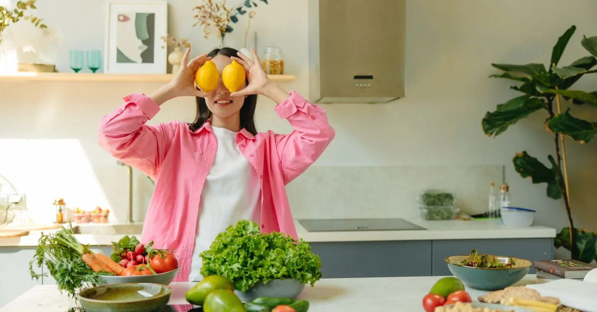 Une femme tenant des citrons de façon ludique devant ses yeux dans une cuisine lumineuse et animée, remplie de légumes frais.